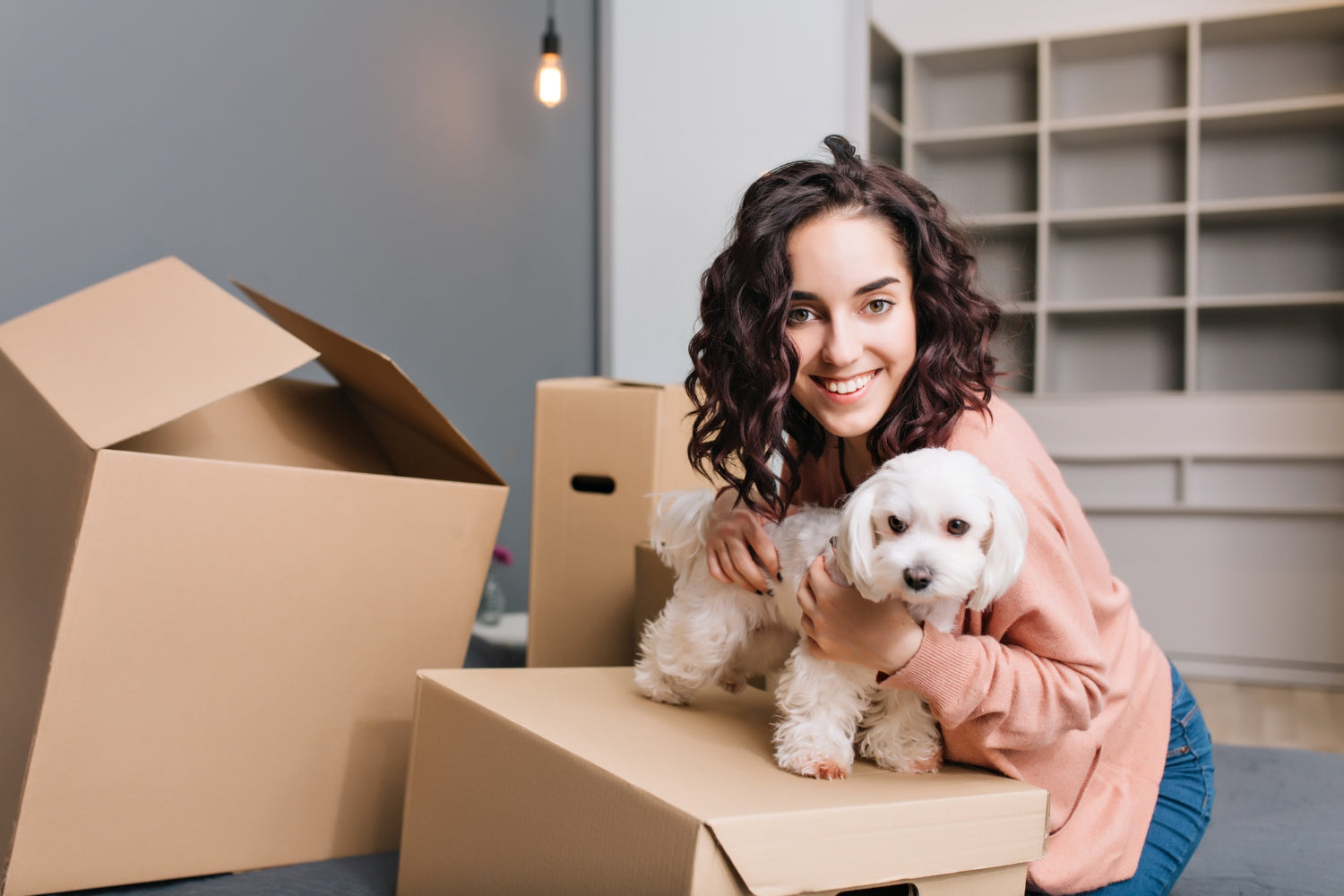 A woman sits beside several cardboard boxes while holding a small white dog, suggesting a home moving or packing scene.